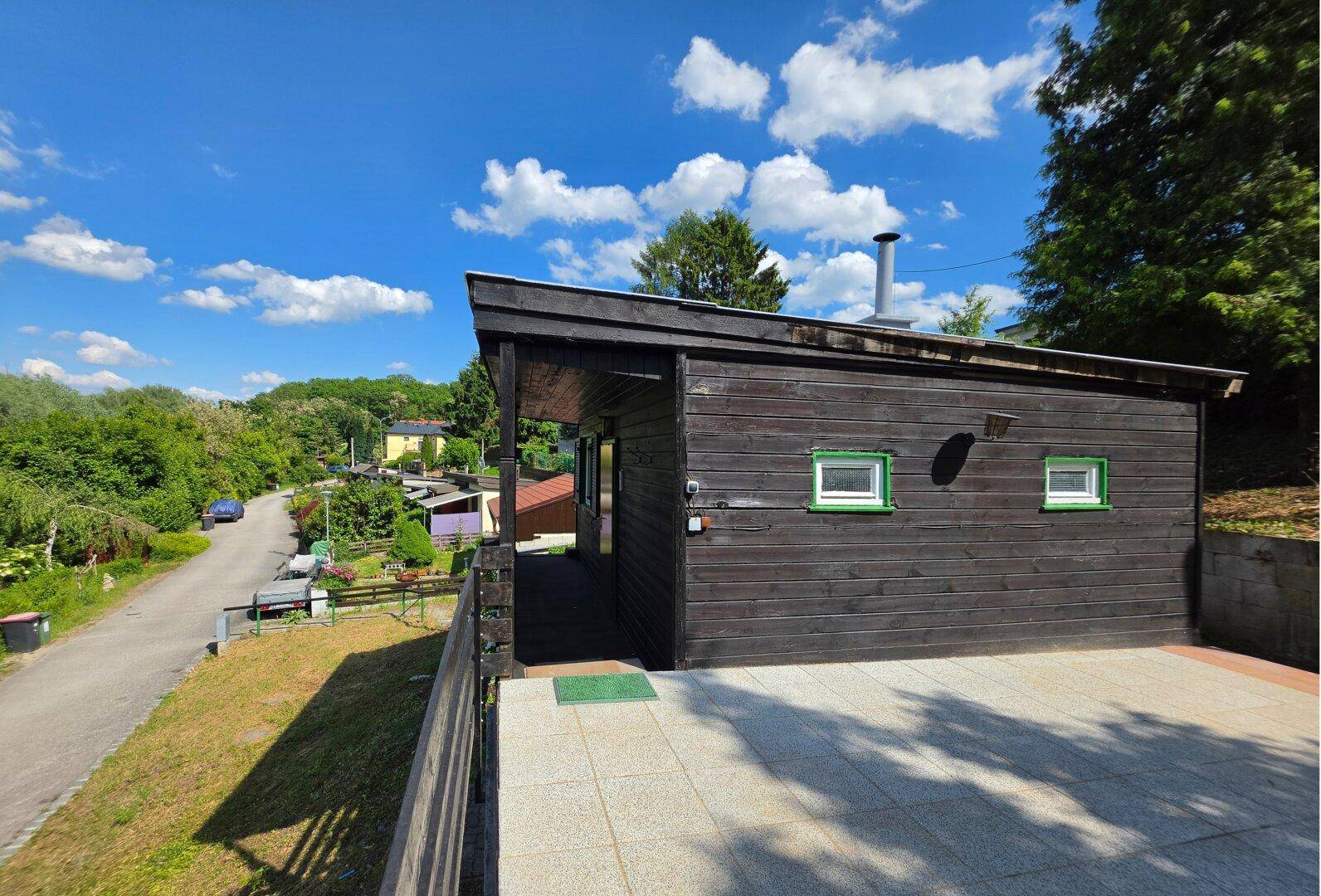 Seitlicher Blick auf das Holzhaus mit großer gepflasterter Terrasse und Blick ins Grüne.