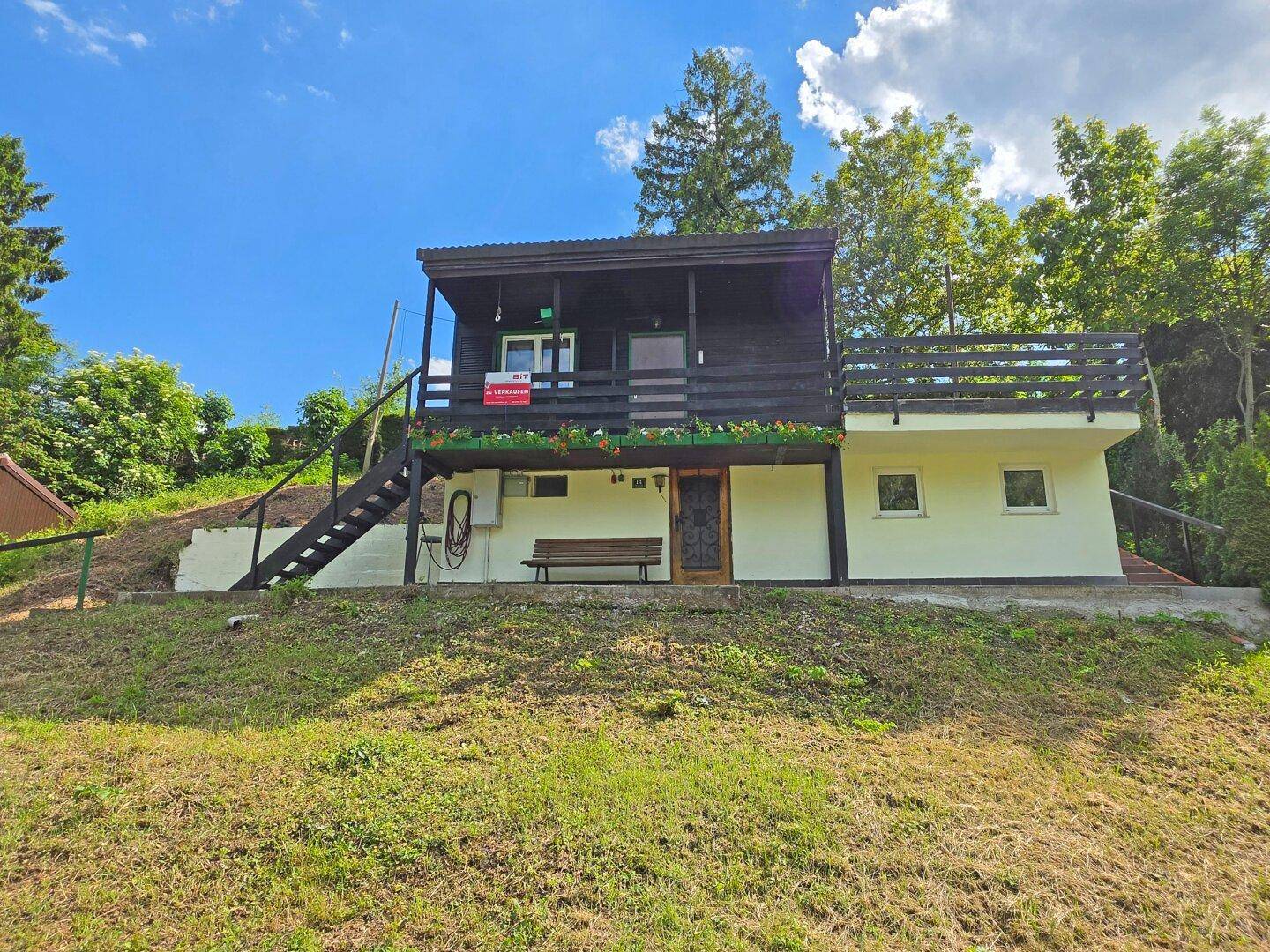 Charmantes Holzhaus mit Balkon und weißem Sockel auf einem sonnigen Hanggrundstück.