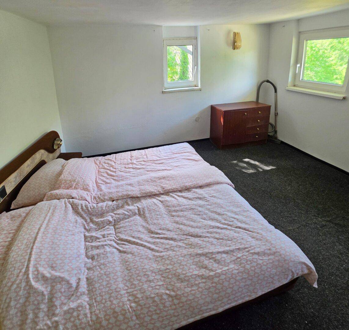 Helles Schlafzimmer mit Doppelbett, Teppichboden und zwei Fenstern mit Blick ins Grüne.
