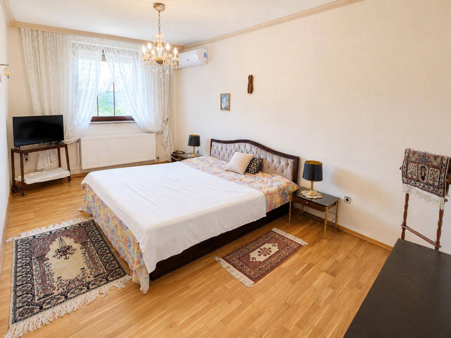 Elegantes Schlafzimmer mit Holzboden, großem Bett, Kronleuchter und Klimaanlage.