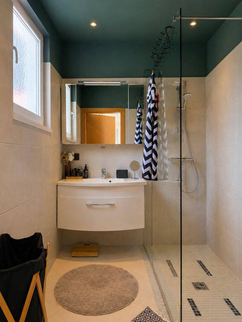 Helles Badezimmer mit Duschkabine, Waschbecken und Fenster, mit einer dunkelgrünen Decke.