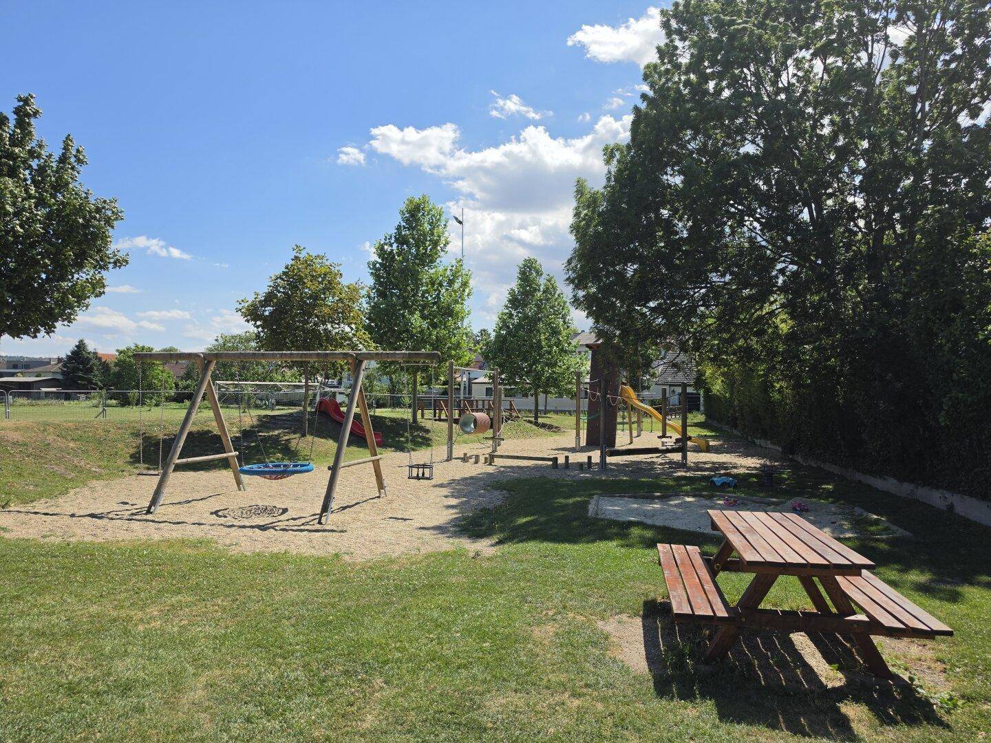 Kinderspielplatz mit Schaukeln, Rutschen und einem Picknicktisch unter Bäumen.