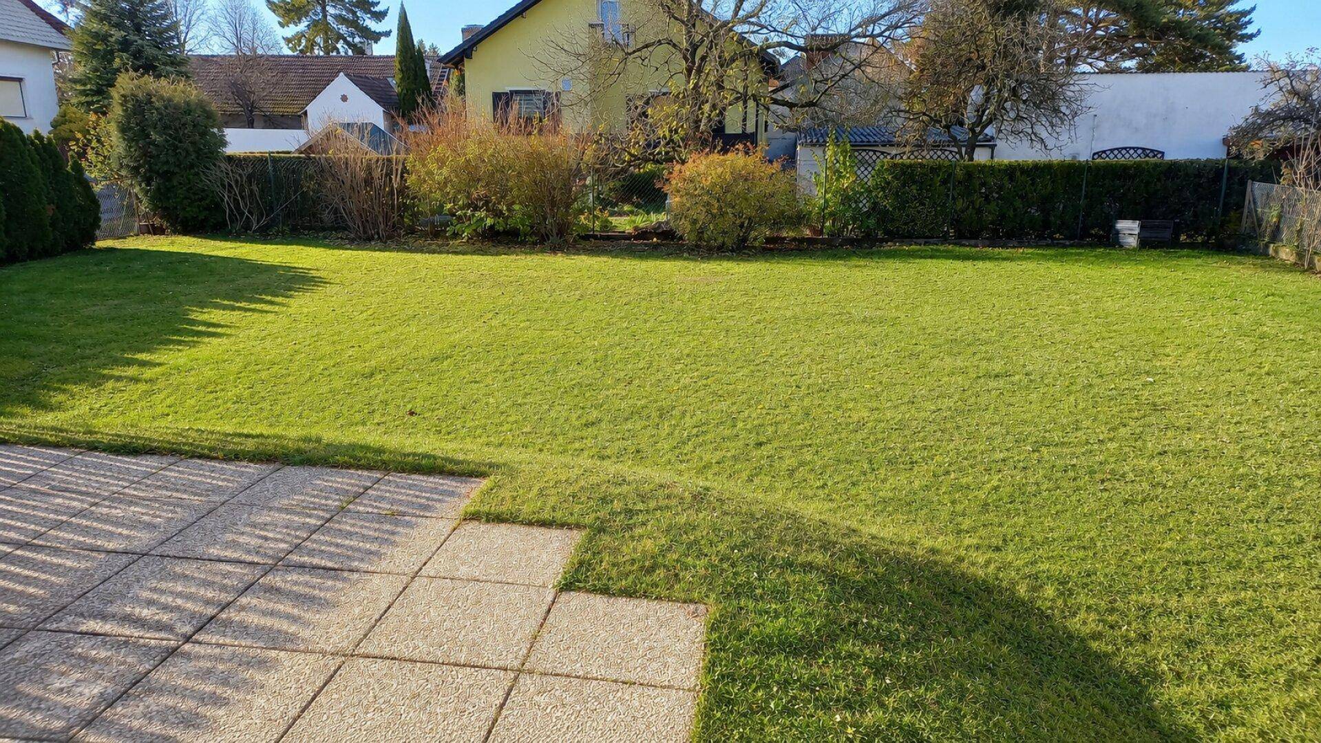 Großer, gepflegter Garten mit Rasenfläche und kleiner Terrasse im Sonnenlicht.