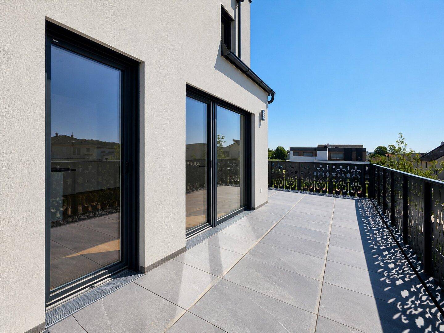 Große, sonnige Terrasse mit Fliesenboden und dekorativem Geländer an einem Neubau.