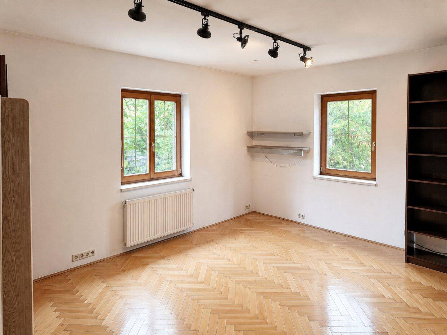 Leeres Zimmer mit Fischgrätparkett, zwei Fenstern und einem Heizkörper, ideal als Schlafzimmer.