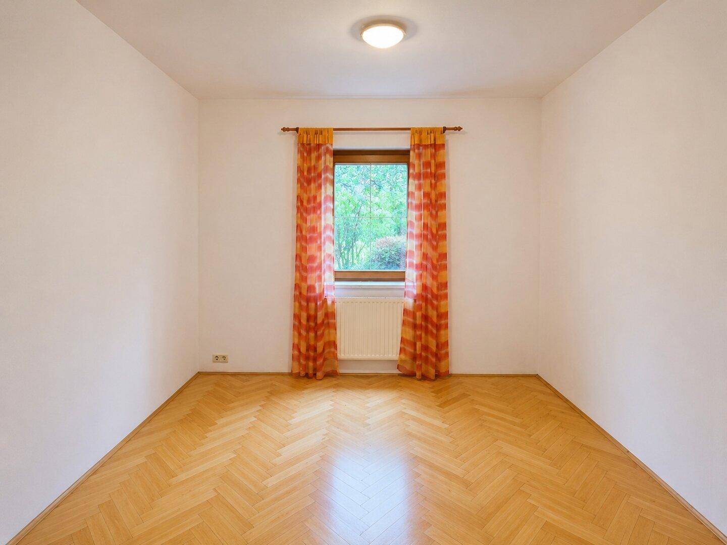 Helles Zimmer mit Fischgrätparkett, einem Fenster mit Vorhängen und einem Heizkörper.