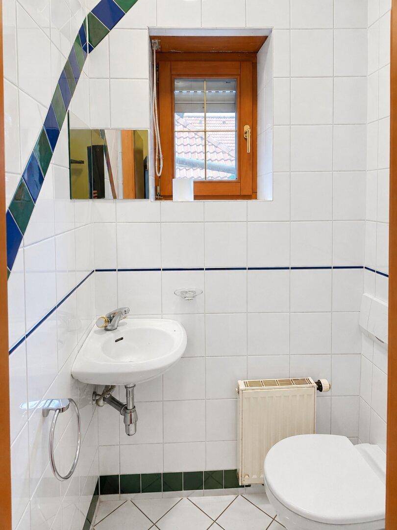 Kleines Gäste-WC mit weißen Fliesen, Waschbecken, Toilette und einem Fenster.