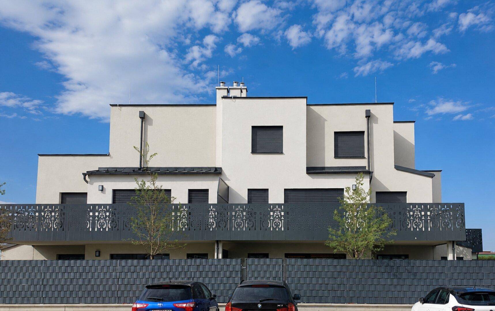 Moderne Wohnanlage mit klaren Linien, Balkonen und einer dekorativen Zaunanlage unter blauem Himmel.