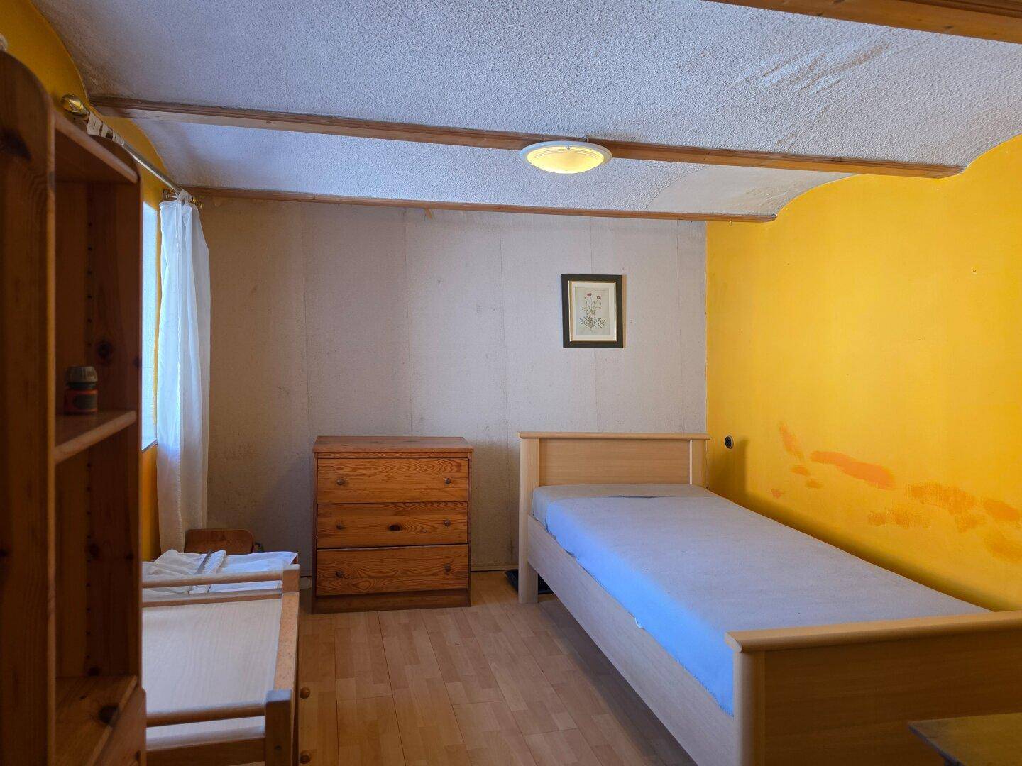 Schlafzimmer mit Holzboden, gelber Akzentwand und sichtbaren Deckenbalken.