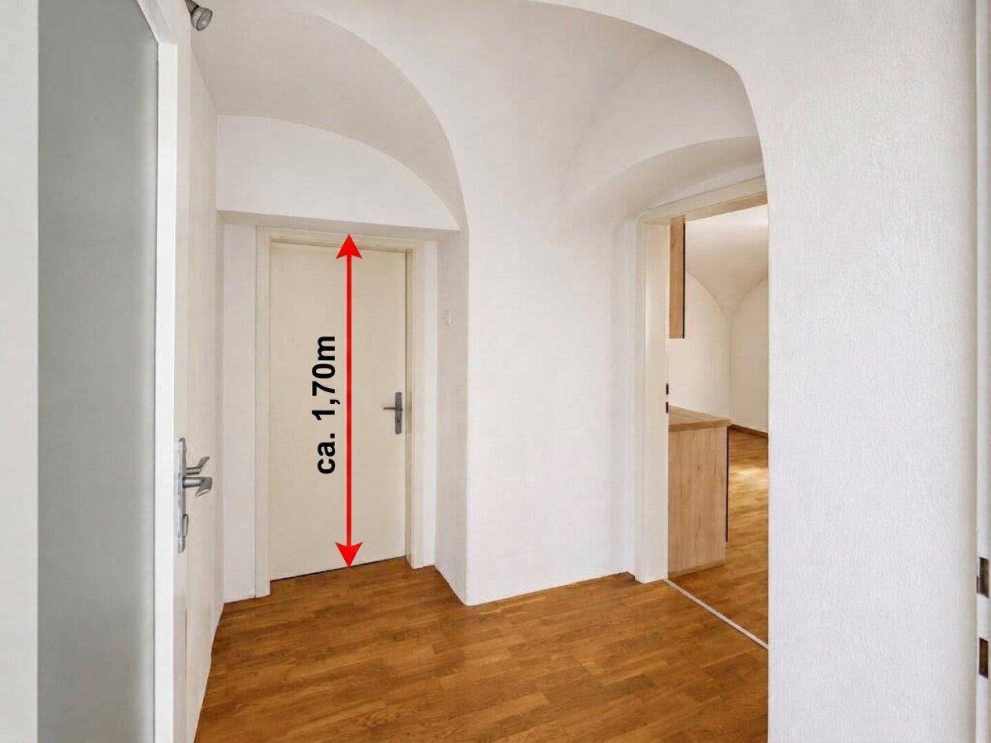 Heller Flur mit gewölbter Decke und Holzboden, gekennzeichnet durch eine niedrige Tür von ca. 1,70m Höhe.