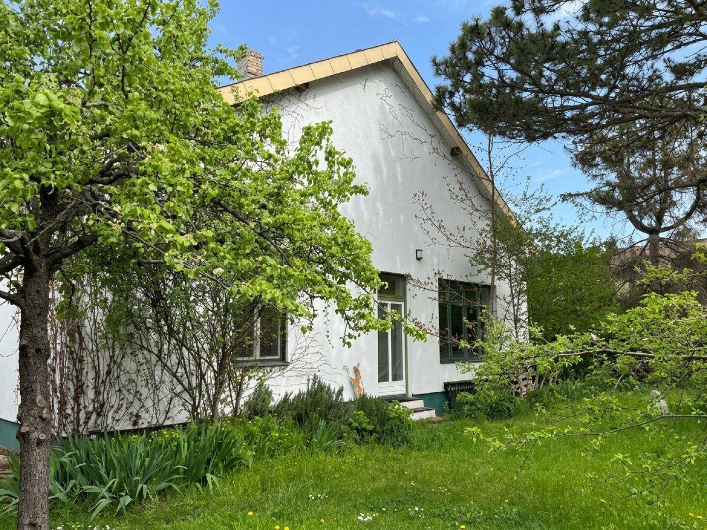 Weißes Wohnhaus mit grüner Umgebung und Bäumen unter einem hellblauen Frühlingshimmel.