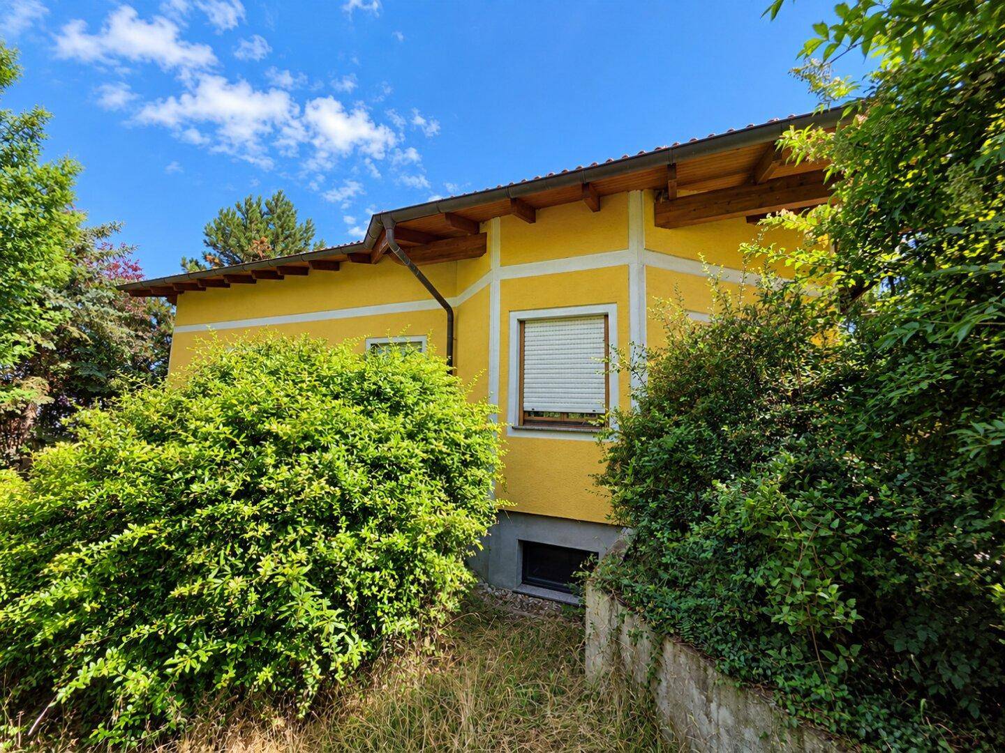 Gelbes Haus mit grauer Sockelwand, umgeben von üppigem Grün unter blauem Himmel.