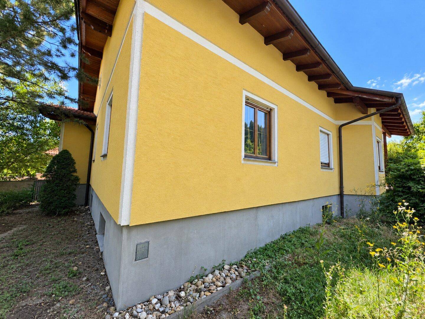 Seitenansicht des gelben Hauses mit grauer Sockelwand und gepflegter Umgebung.