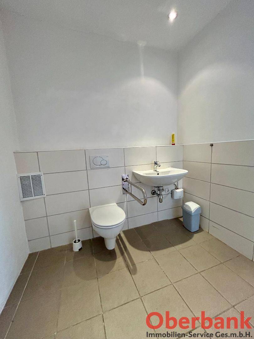 Barrierefreies WC mit Haltegriffen, wandmontierter Toilette und Waschbecken in hellen Fliesen, gut belüftet.