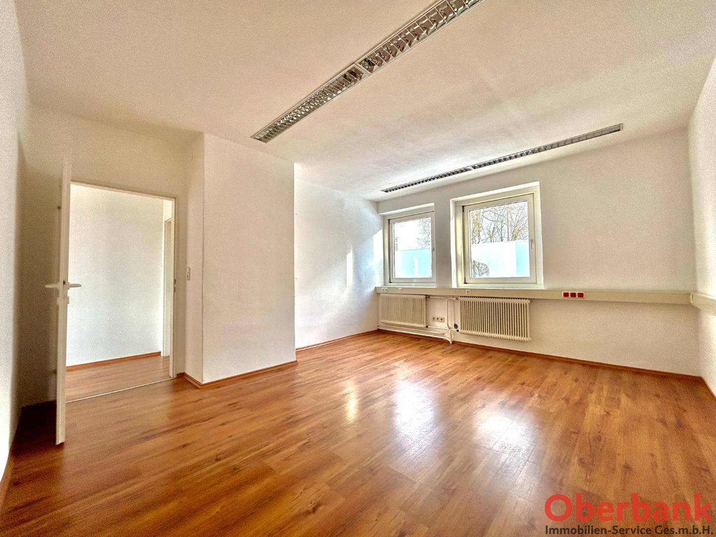 Geräumiges Büro mit Laminatboden, weißen Wänden und zwei Fenstern, bietet eine ruhige Arbeitsumgebung.