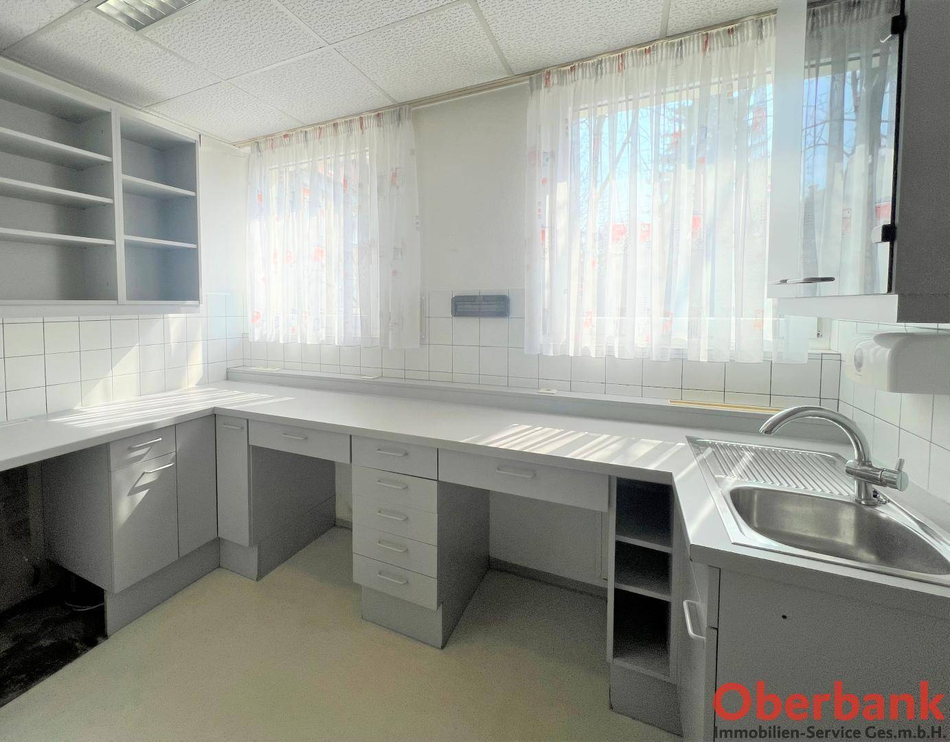 Helle Küche mit weißen Schränken, Arbeitsflächen und einem Spülbecken unter den Fenstern.