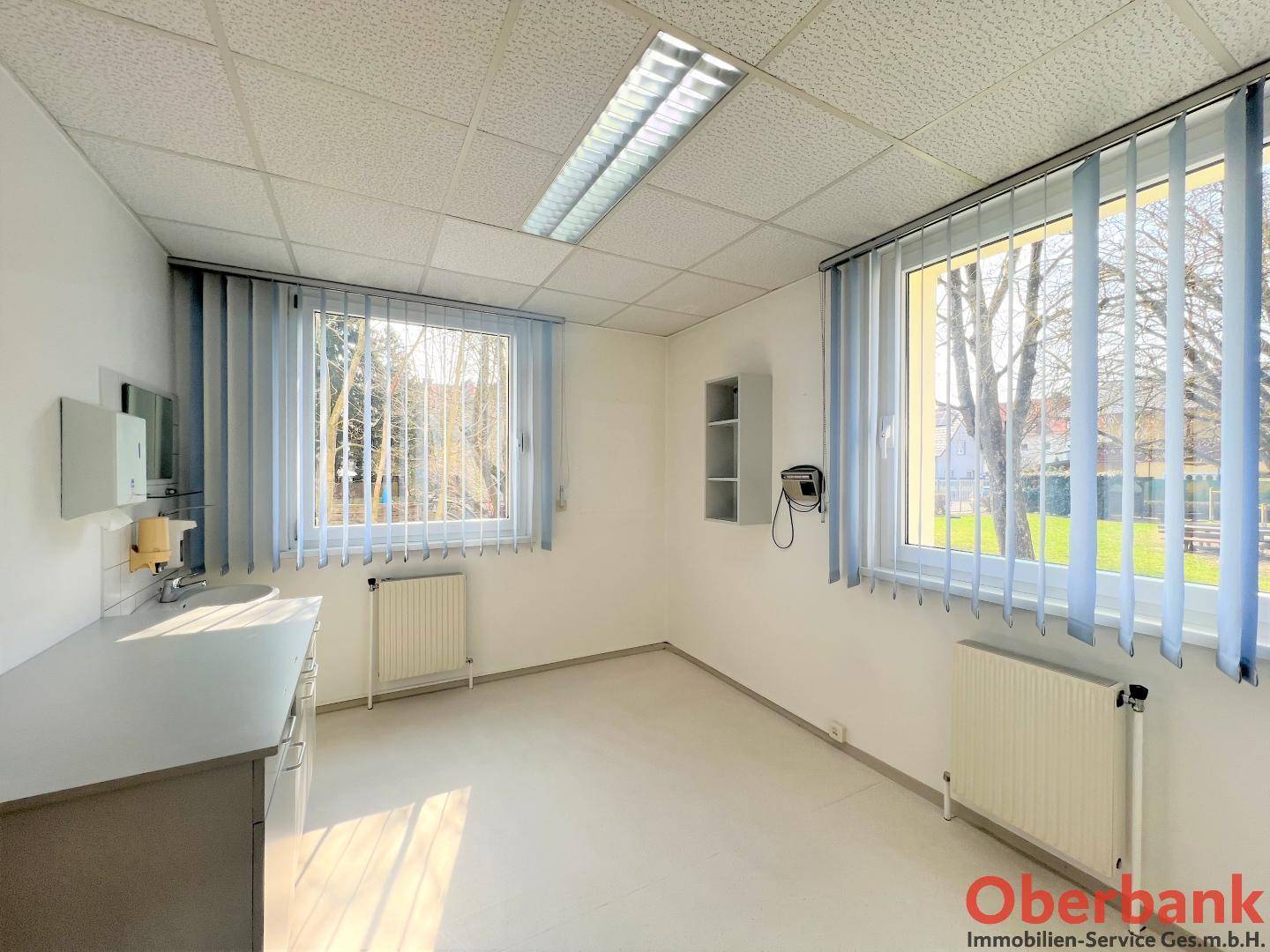 Helles Büro mit zwei großen Fenstern, Heizkörpern und einem Waschbecken an der Wand.