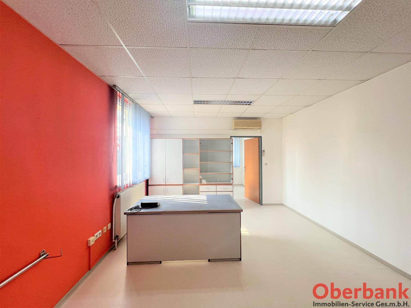 Geräumiges Büro mit roter Akzentwand, Klimaanlage und Blick auf einen weiteren Raum.