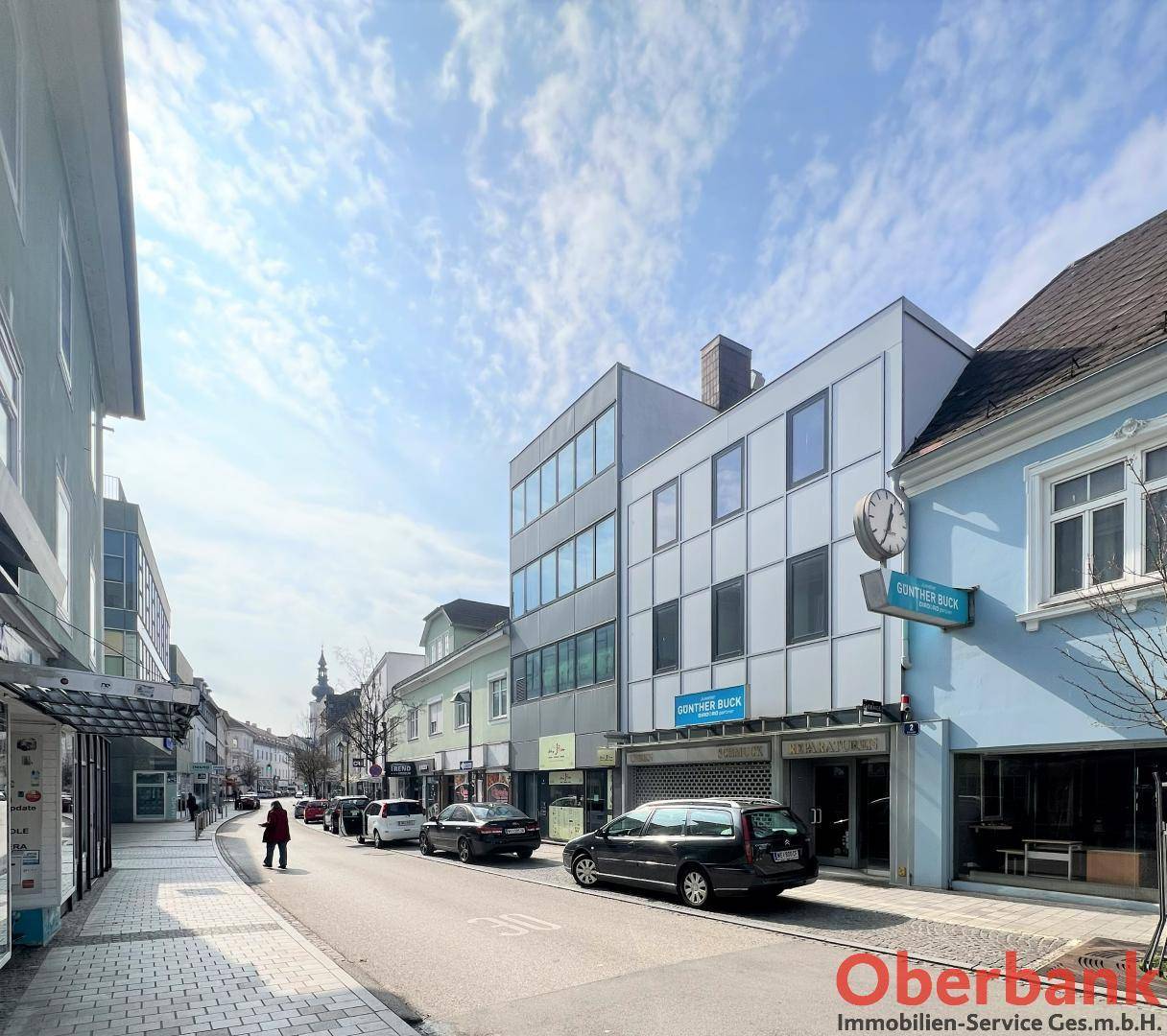 Belebtes Stadtbild mit Geschäften und Gebäuden entlang einer Straße unter blauem Himmel.