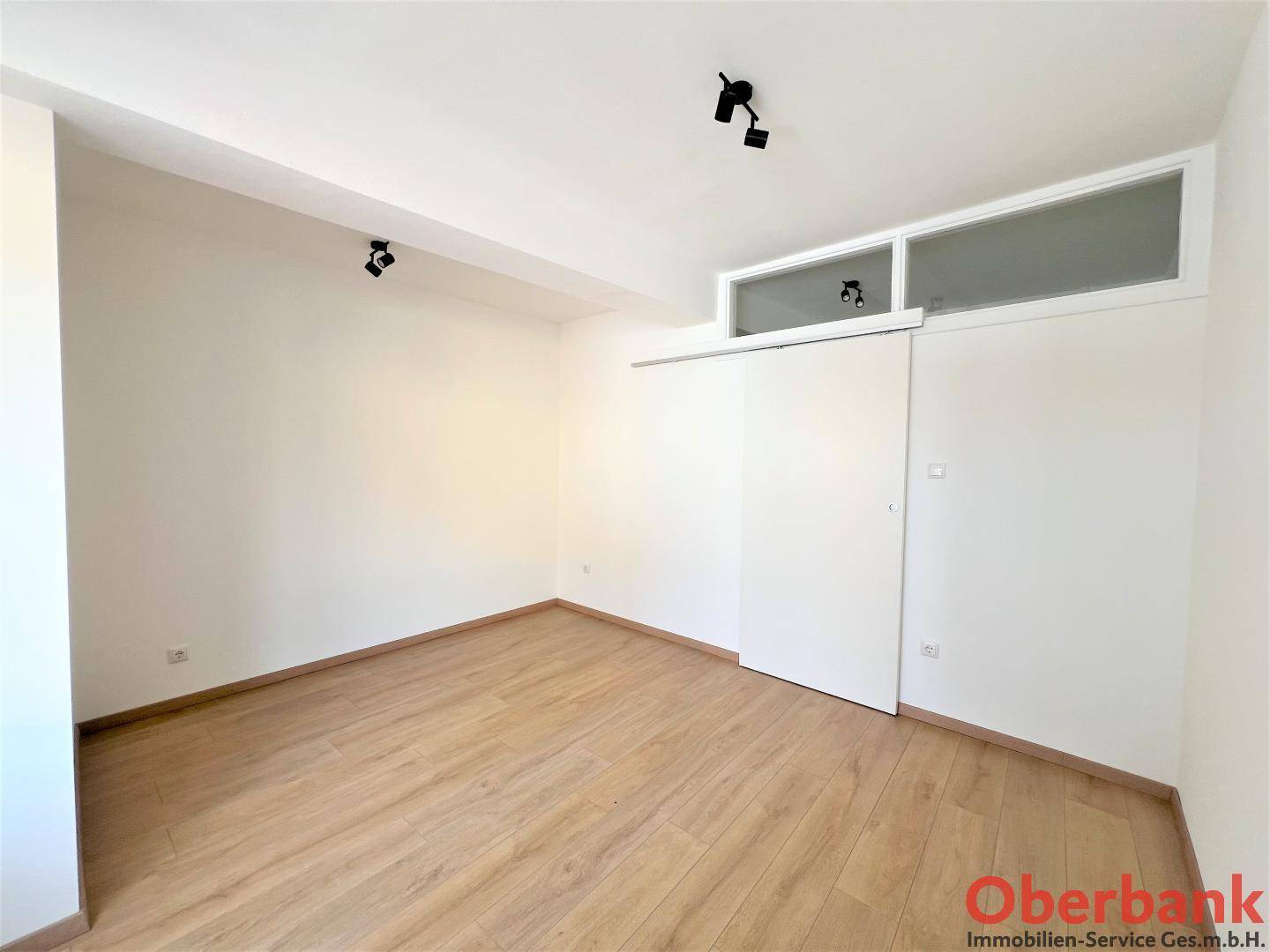 Helles Zimmer mit Laminatboden und einer Schiebetür, die zu einem weiteren Raum führt.