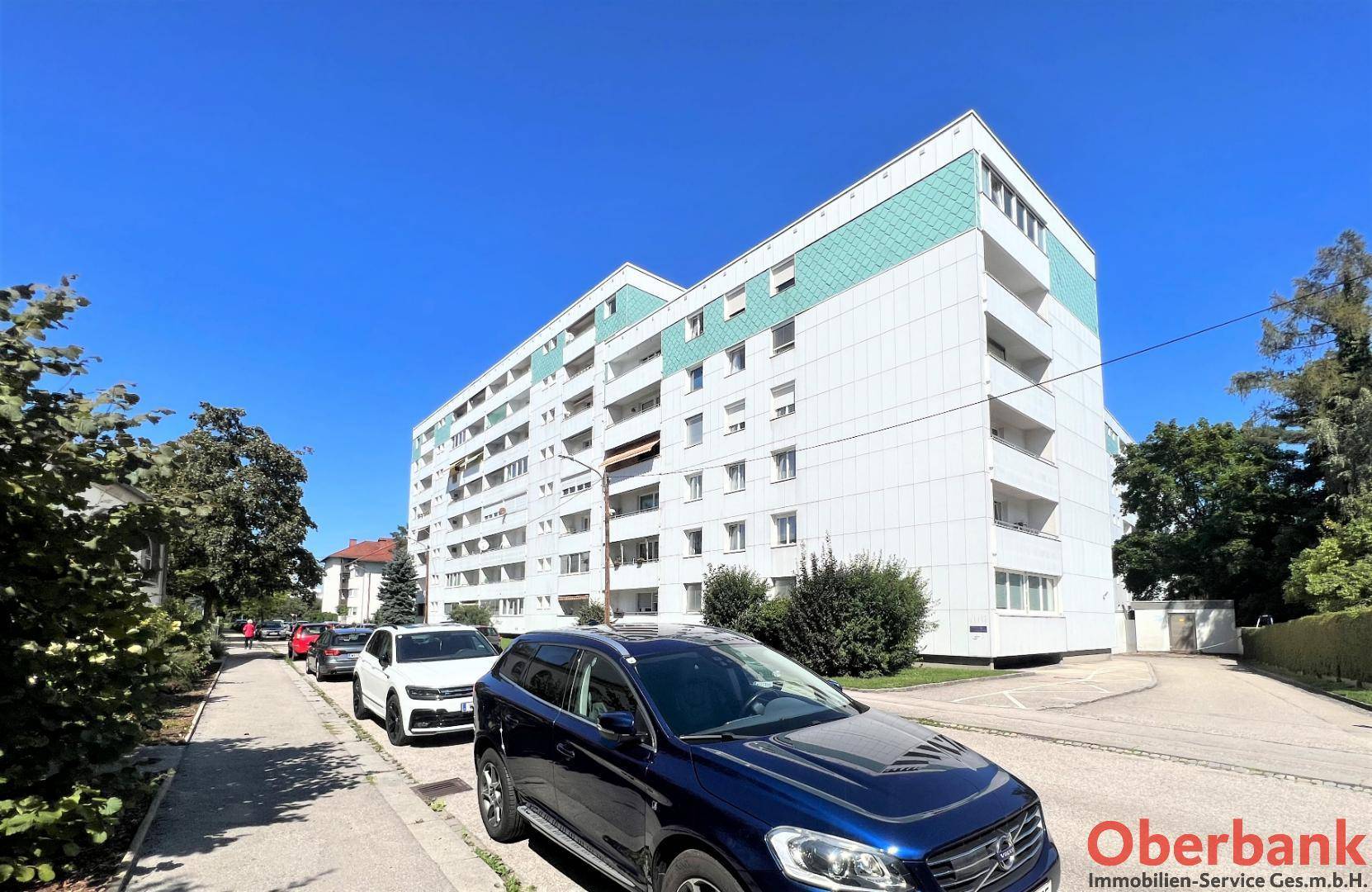 Großes Wohngebäude mit weißer und türkiser Fassade, umgeben von Bäumen und parkenden Autos.