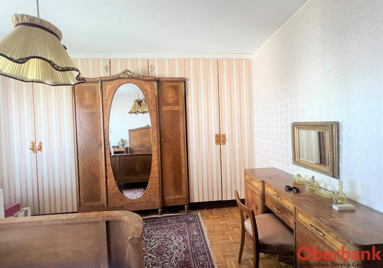 Schlafzimmer mit klassischem Holzschrank, Spiegel und Kommode, ergänzt durch gemusterte Tapeten.