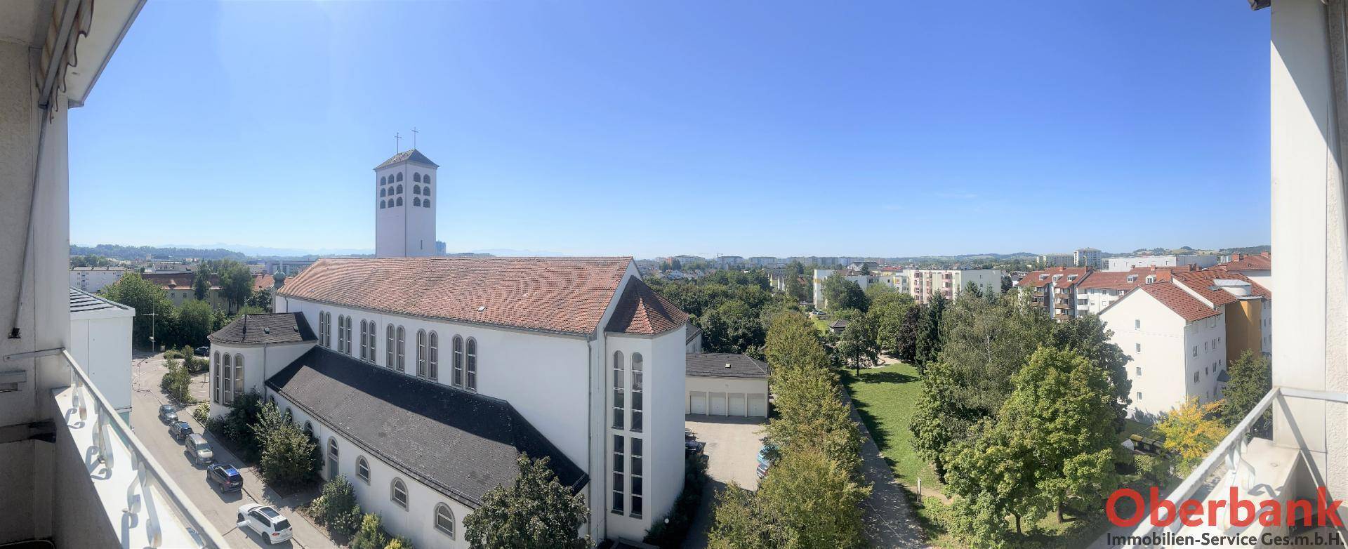 Weitläufiger Panoramablick von einem Balkon auf eine Kirche und die grüne Stadtlandschaft.