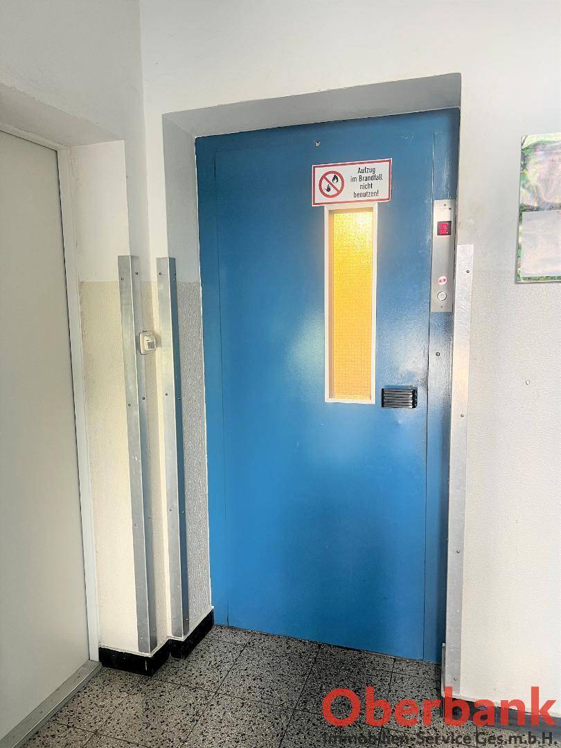 Blauer Aufzug in einem Flur mit gefliestem Boden und Hinweisschildern.