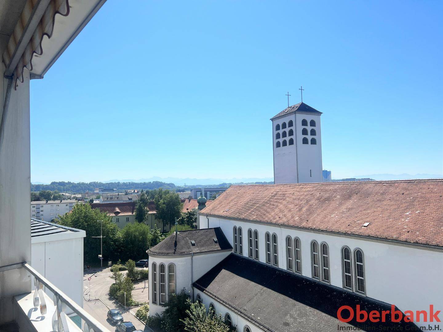 Panoramablick von einem Balkon auf eine Kirche und die umliegende Stadtlandschaft mit Bergen im Hintergrund.