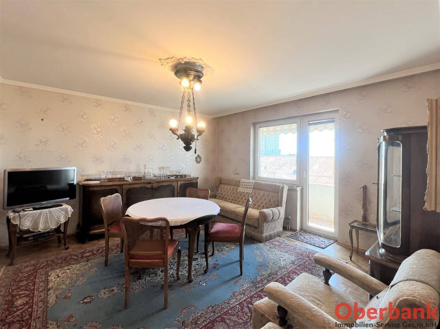 Gemütlicher Wohn-Essbereich mit traditionellen Möbeln, Teppich und Zugang zu einem Balkon.