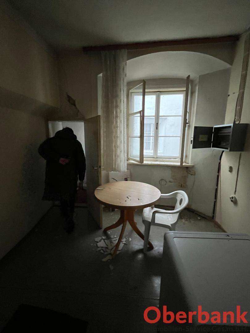 Dunkler Raum mit offenem Fenster, Tisch und Stuhl, der deutlichen Renovierungsbedarf aufweist.