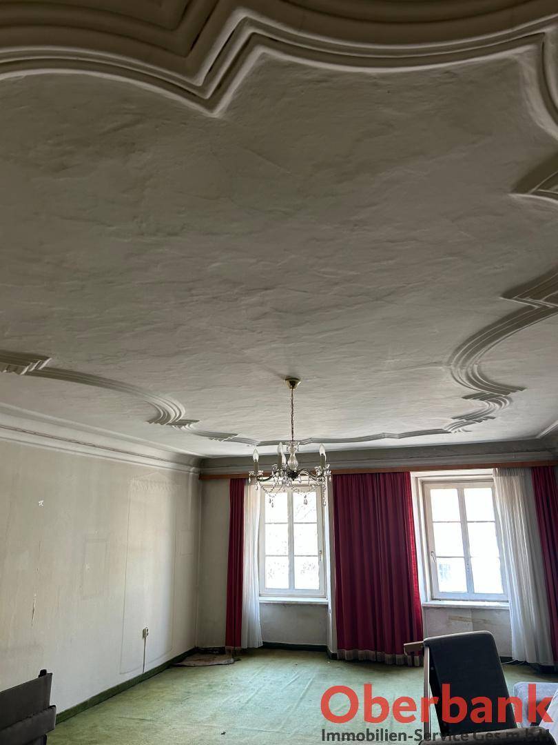 Zimmer mit aufwendiger Stuckdecke, Kronleuchter, grünem Teppich und roten Vorhängen an den Fenstern.
