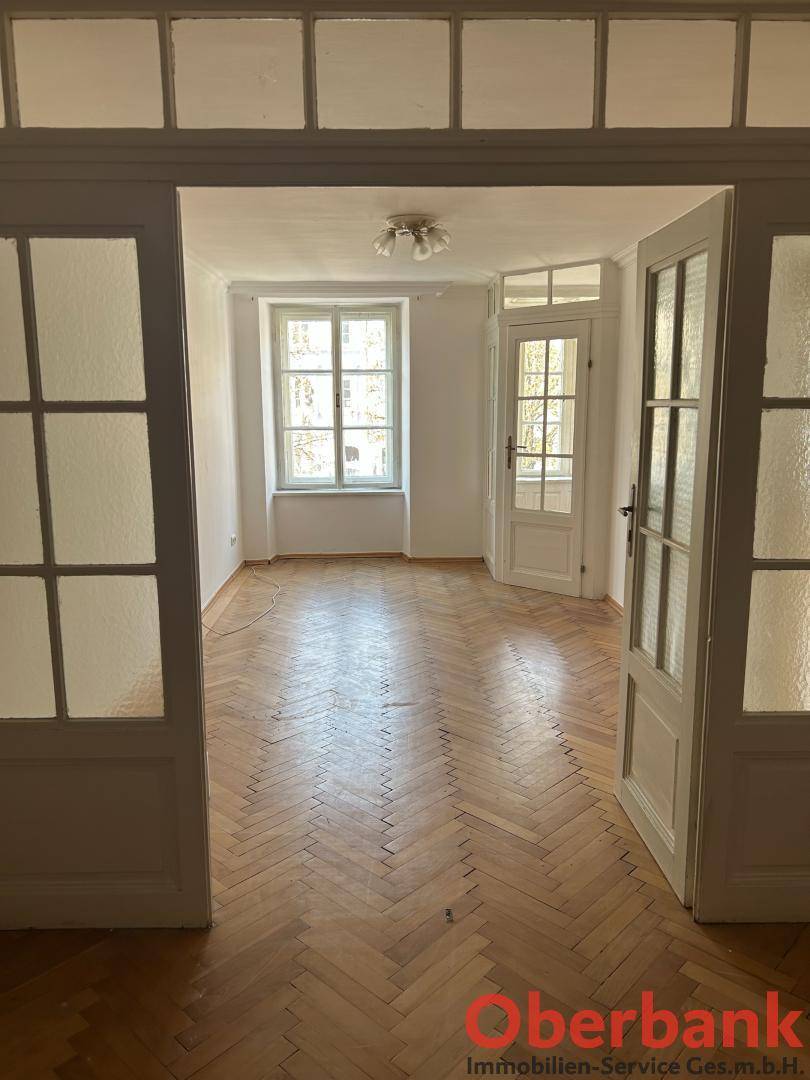 Leeres Zimmer mit Fischgrätparkett, weißen Wänden und großen Fenstern, zugänglich durch Flügeltüren mit Glaseinsätzen.
