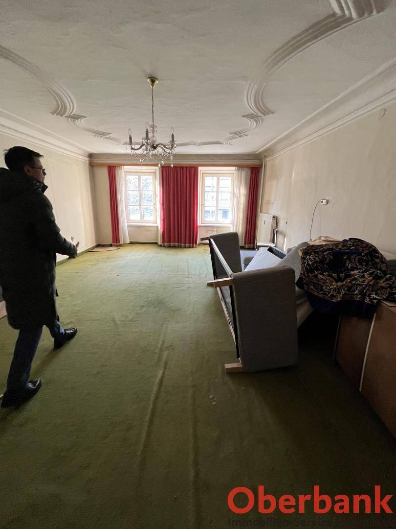 Geräumiges Wohnzimmer mit grünem Teppichboden, Stuckdecke und zwei hohen Fenstern.
