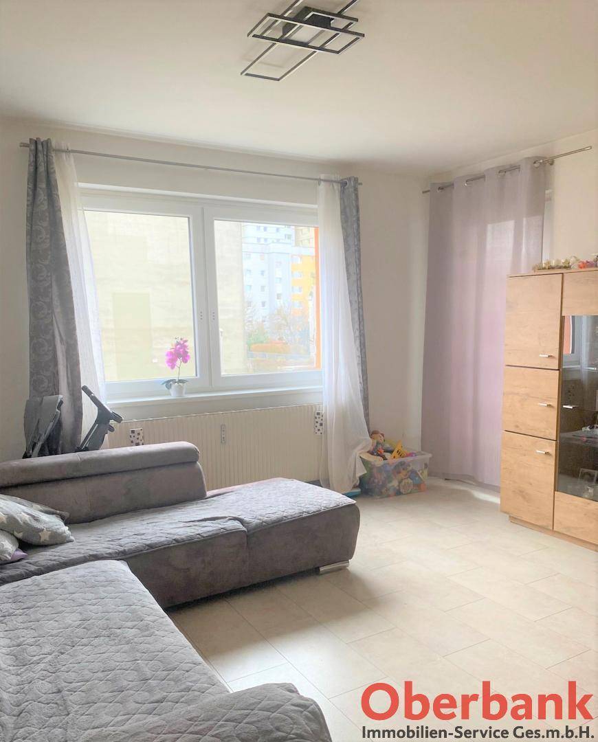 Helles Wohnzimmer mit großem Fenster, beigem Sofa und Holzschrank, freundlich eingerichtet.