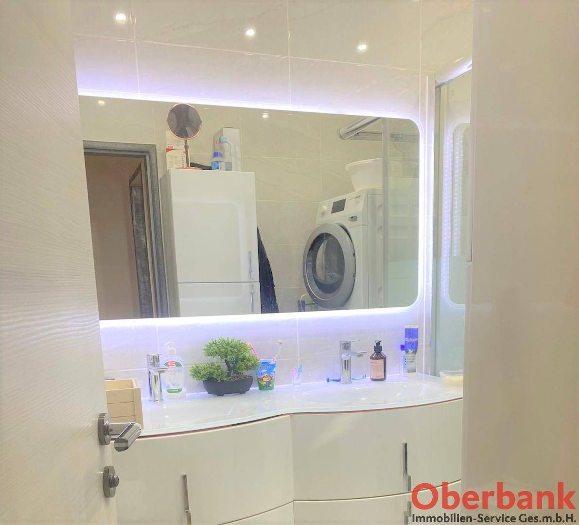 Modernes Badezimmer mit beleuchtetem Spiegel, Waschbeckenunterschrank und Waschmaschine im Hintergrund.