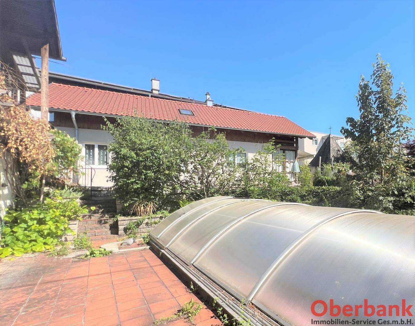 Gartenbereich mit überdachtem Pool und gepflasterter Fläche, umgeben von Grün.