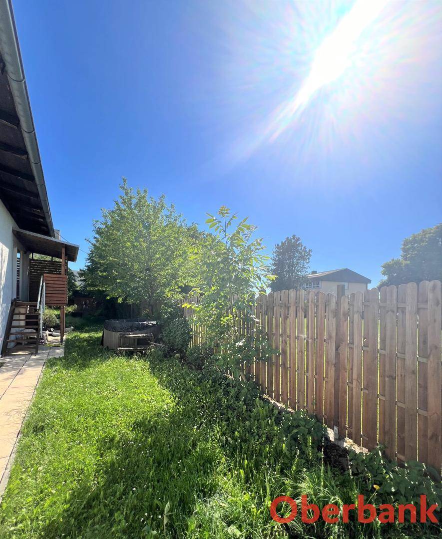Seitlicher Gartenbereich mit Rasen, Holzzaun und jungen Bäumen unter blauem Himmel.