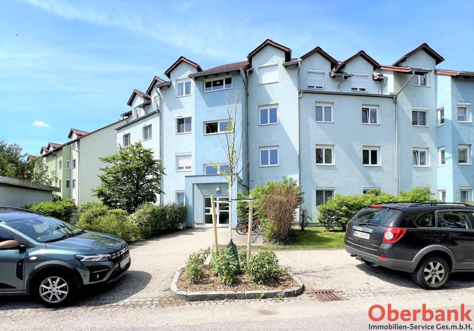 Ansicht des Wohngebäudes mit hellblauer Fassade, Eingangsbereich und gepflegter Außenanlage.
