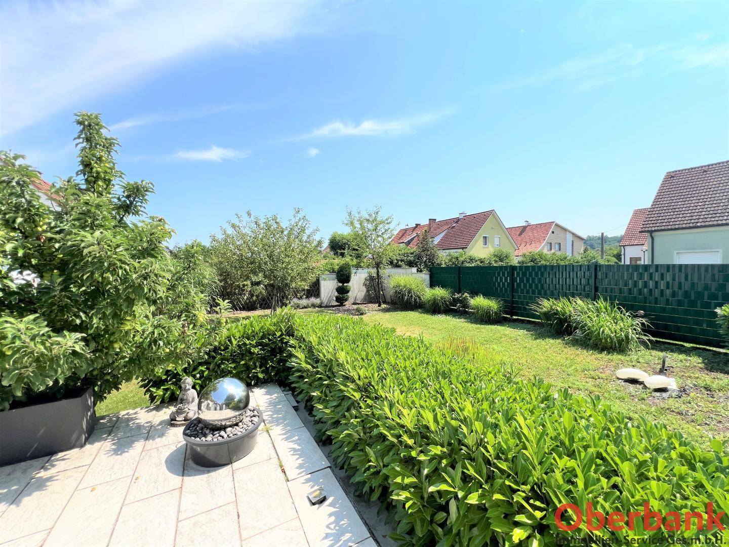 Gepflegter Gartenbereich mit Rasen, Sträuchern, einem kleinen Brunnen und Blick auf Nachbarhäuser.
