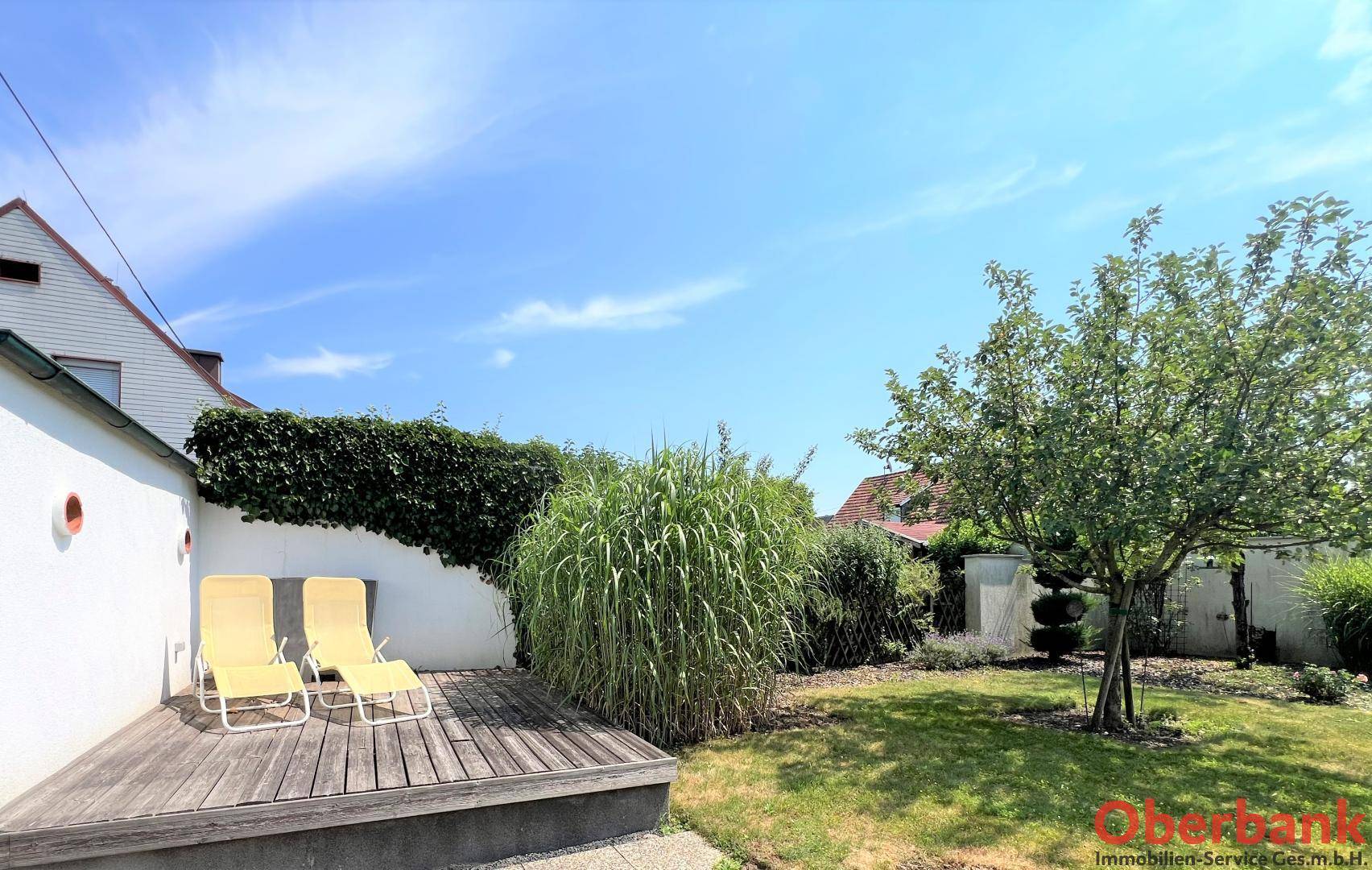 Sonniger Gartenbereich mit Holzdeck, Liegestühlen und üppiger Bepflanzung unter blauem Himmel.