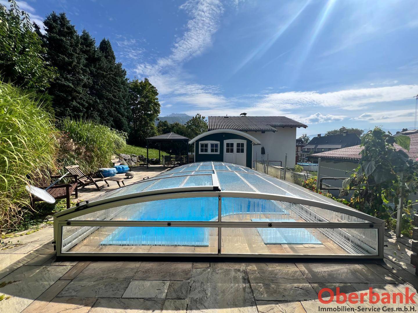 Überdachter Swimmingpool im Freien, umgeben von einer Steinterrasse und grüner Umgebung.