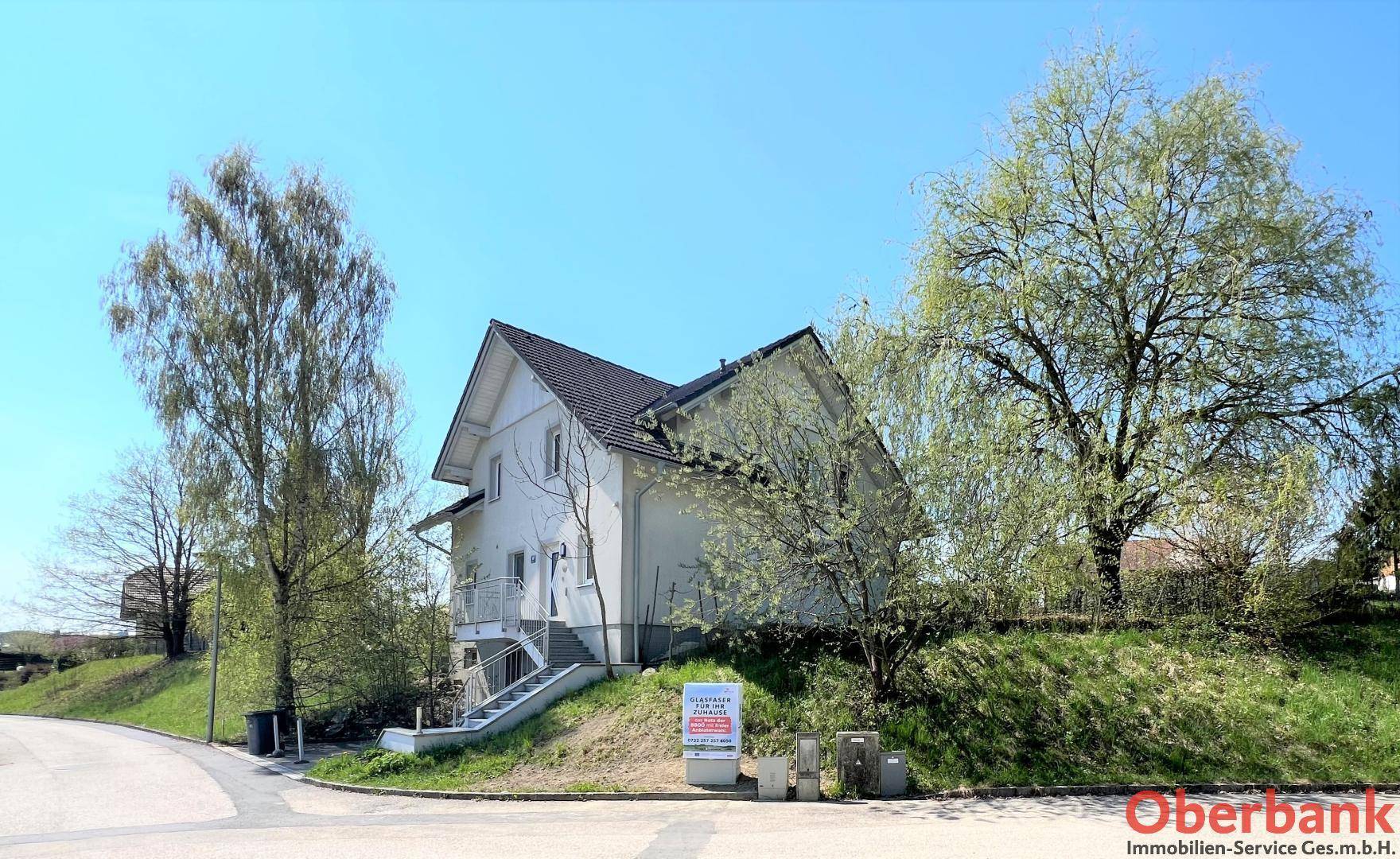 Gesamtansicht des Hauses von der Straßenecke, eingebettet in eine grüne Umgebung mit Bäumen und Hügeln.