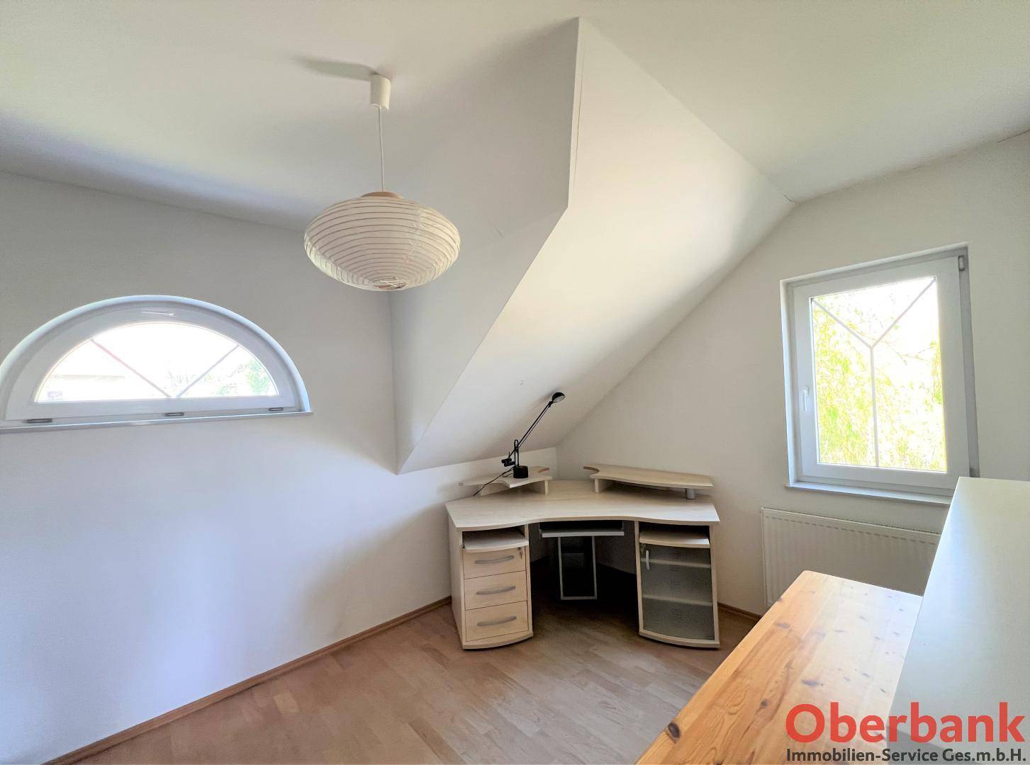 Arbeitszimmer mit Holzboden, Schreibtisch und zwei Fenstern, eines davon rund.