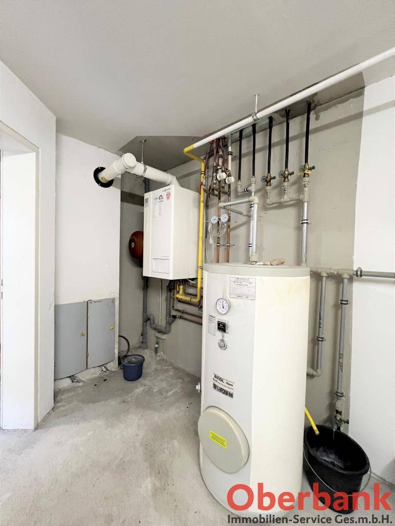 Unfertiger Hauswirtschaftsraum im Keller mit Warmwasserspeicher, Rohren und Elektroinstallationen.