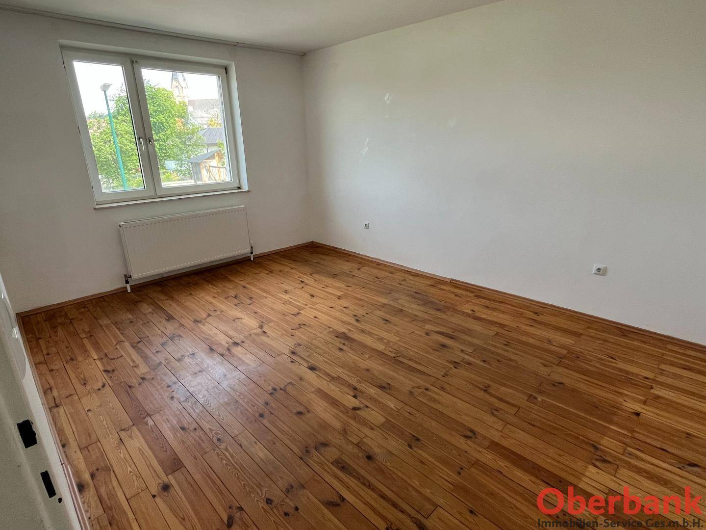 Leeres Zimmer mit hellem Holzplankenboden und einem Fenster mit Blick ins Grüne.