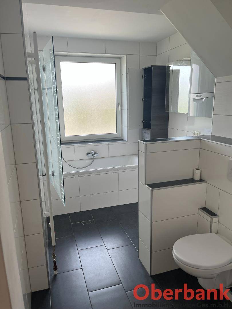 Helles Badezimmer mit Badewanne, Toilette, Fenster und dunklen Bodenfliesen in gutem Zustand.