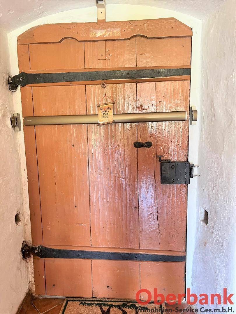 Alte, rustikale Holztür mit schweren Eisenbeschlägen und einem traditionellen Riegel, die zu einem Innenbereich führt.