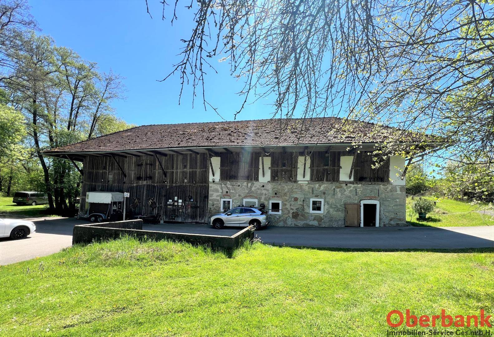 Großes, traditionelles Bauernhaus mit Steinfassade und Holzelementen, umgeben von grüner Landschaft und Parkplätzen.