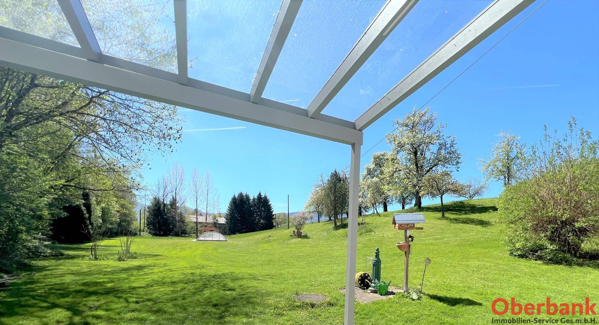 Weitläufiger Blick über die grüne Landschaft von der überdachten Terrasse, mit blühenden Bäumen im Hintergrund.